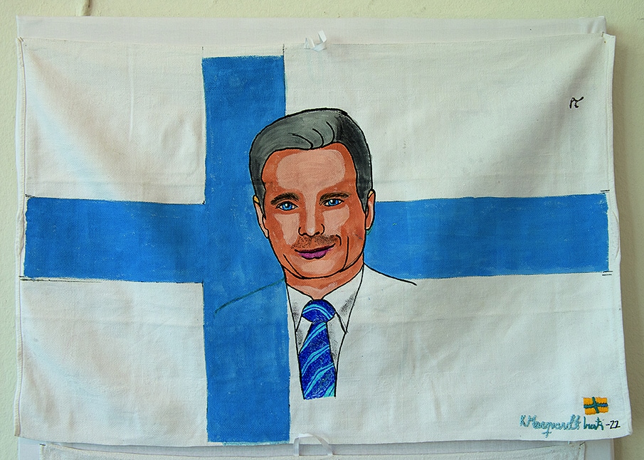 En kökshandduk med blått kors som finska flaggan. I mitten av flaggan porträtt på en välkammad man med slipps som ler.