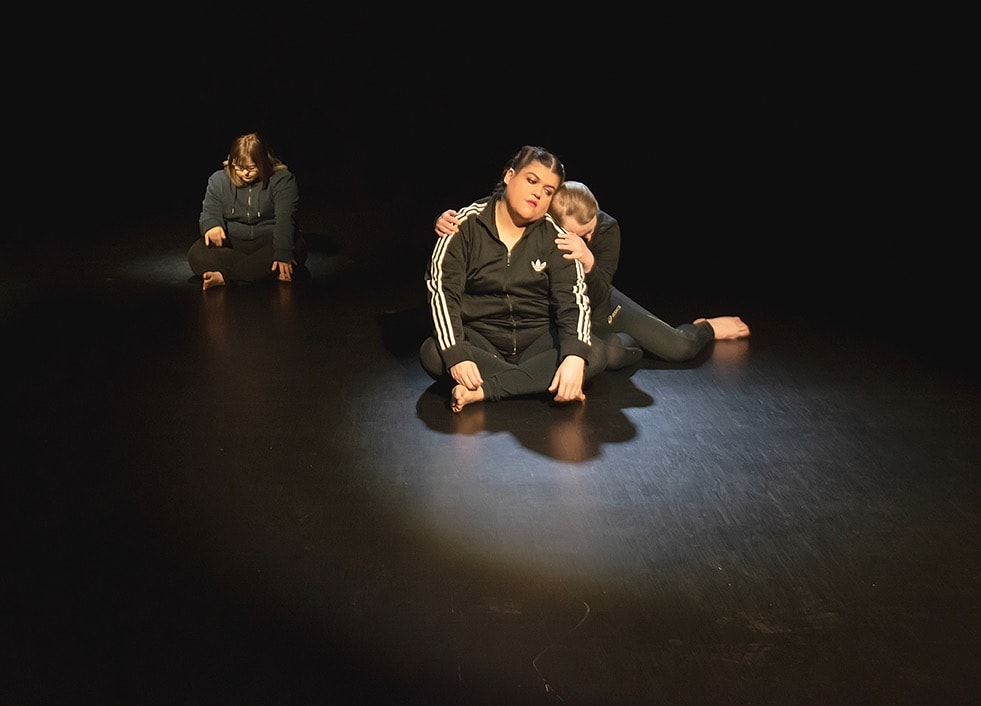 Scenbild. Tre dansare sitter på det svarta scengolvet. Carolina Coreras sitter i skräddarställning. Bakom henne sitter Niklas Hjelmvik som lutar sig mot henne.